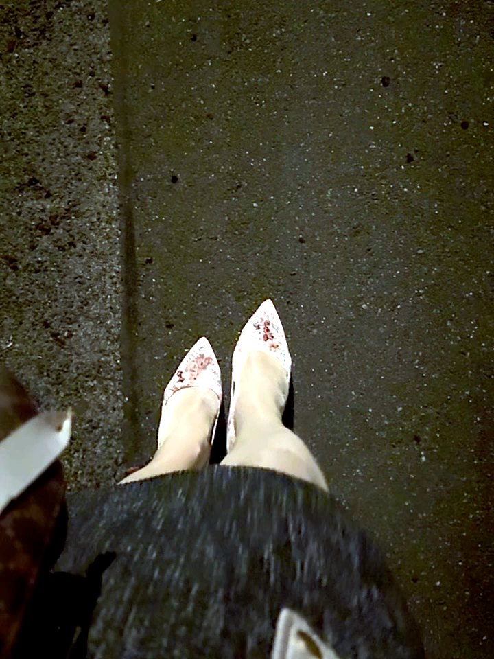Florale Pumps auf dunklem Asphalt neben dem Bordstein, darüber der Saum eines grauen Rocks