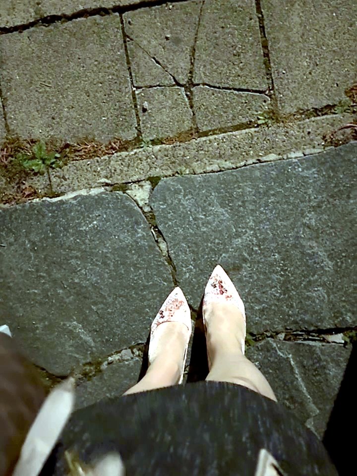 Blick von oben auf florale Pumps, grauen Tweedrock und helle Jacke auf Pflastersteinen