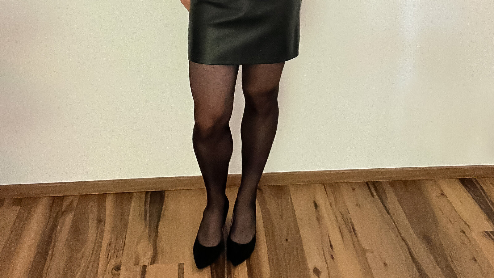 Beine in schwarzer Strumpfhose mit schwarzen Pumps unter einem schwarzen Lederrock