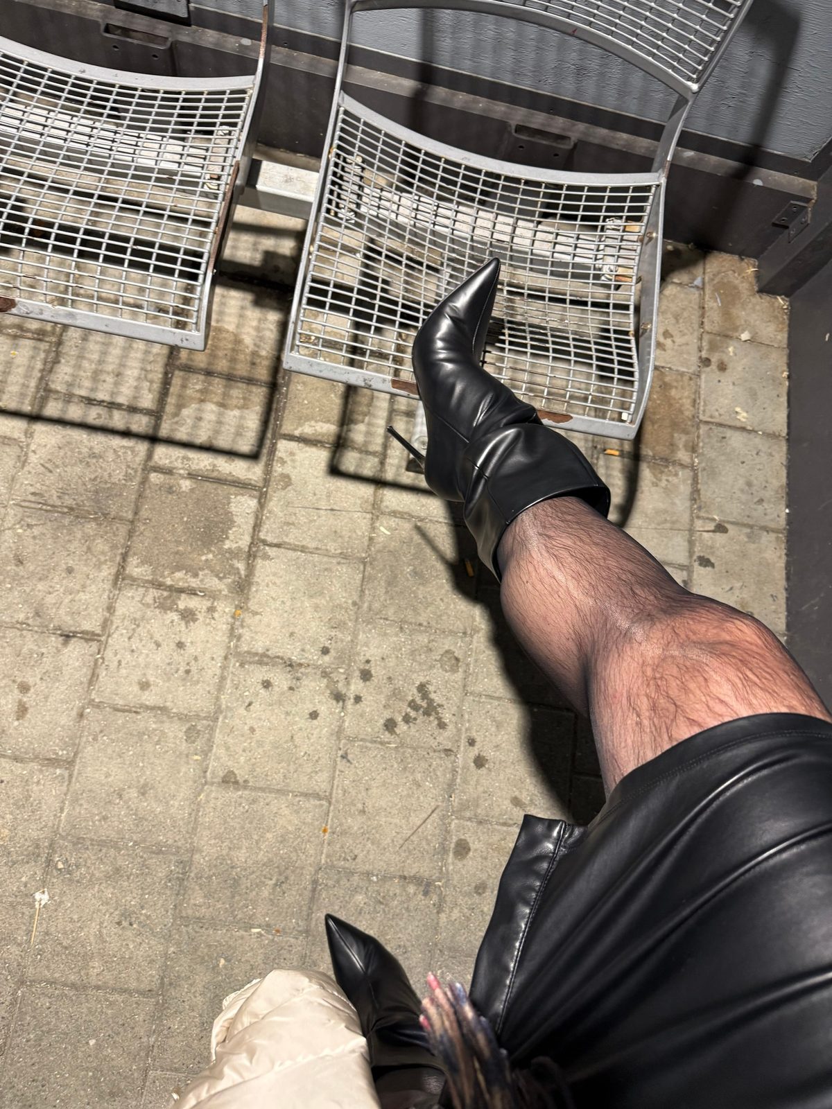Hohe schwarze Stiefel mit Stiletto-Absatz, Bein angehoben vor Metallbänken am Bahnhof