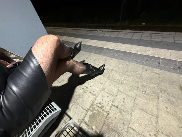 Beine in schwarzer Strumpfhose und spitzen schwarzen Stiefeln, übereinandergeschlagen auf einer Bank am Bahnsteig bei Nacht
